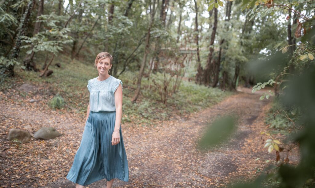 Aurore marche en forêt et sourit à l’objectif, incarnant un postnatal serein et un accompagnement postpartum chaleureux